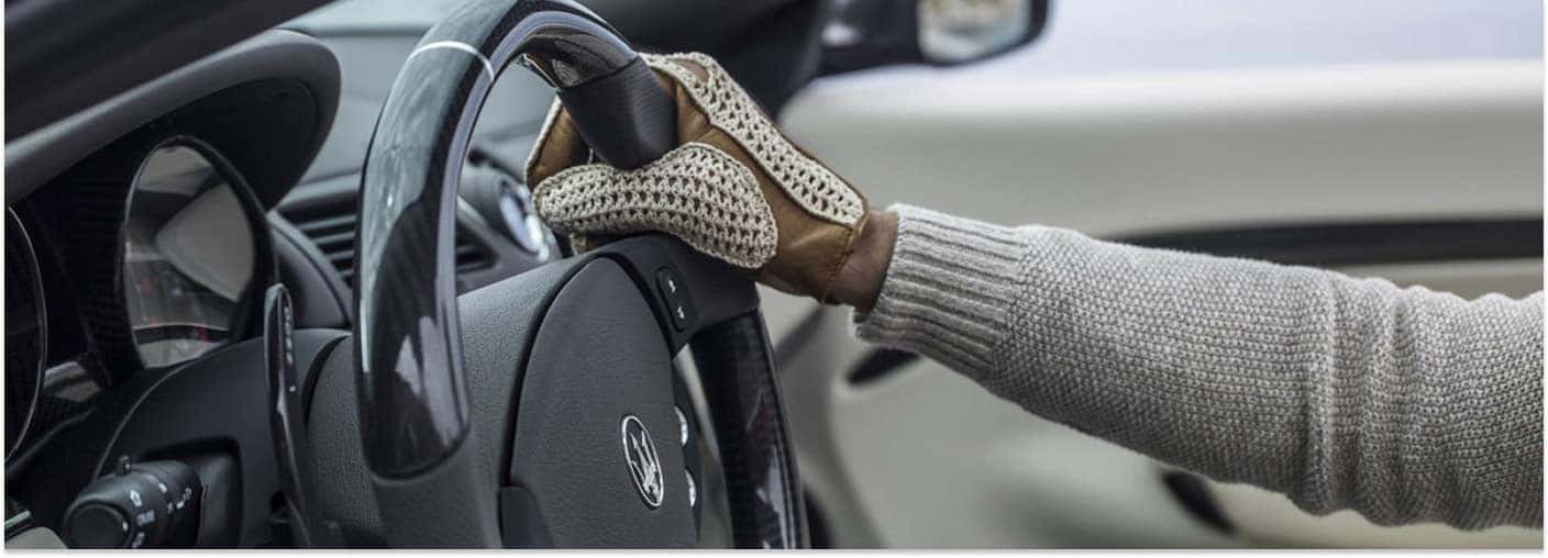 Driver hand in a woven glove resting on a Maserati carbon-fiber steering wheel with a column-mounted paddle shifter.