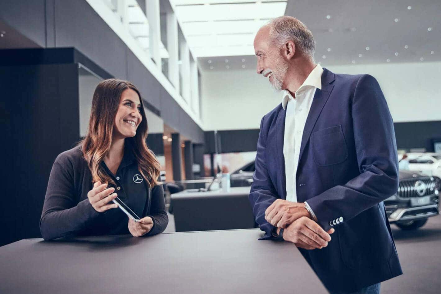 A Mercedes-Benz Sales Associate holds a tablet while assisting a smiling customer inside a dealership.