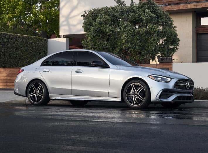 2026 Mercedes-Benz C-Class in Cirrus Silver Metallic, side profile parked outside a modern home on a wet street.