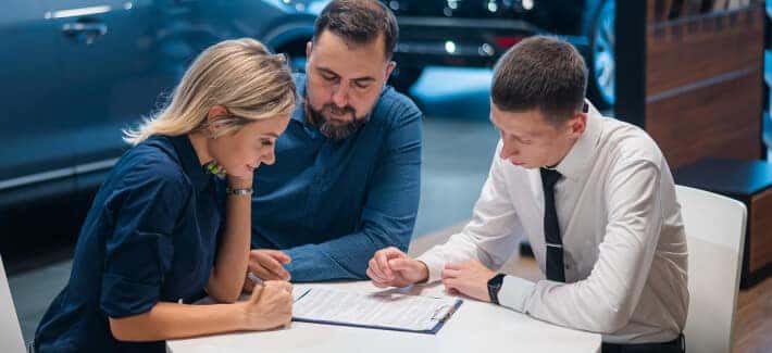 Finance advisor reviews paperwork with two customers at a dealership table, with a luxury vehicle behind them.