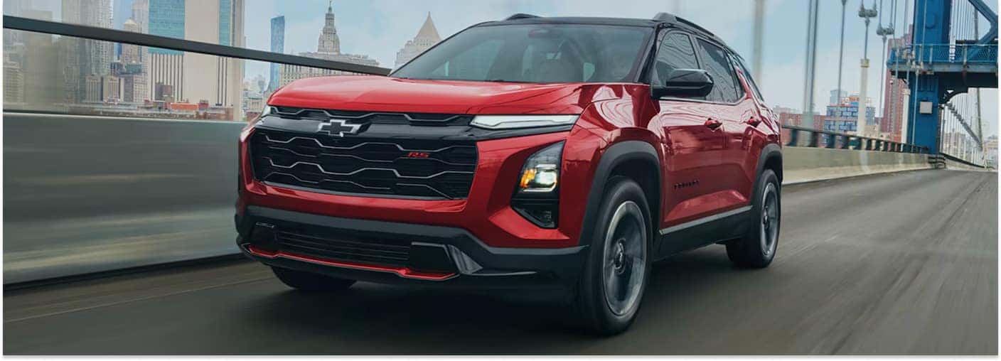 Front three-quarter view of a red 2027 Chevrolet Equinox RS driving across a bridge with a city skyline background.
