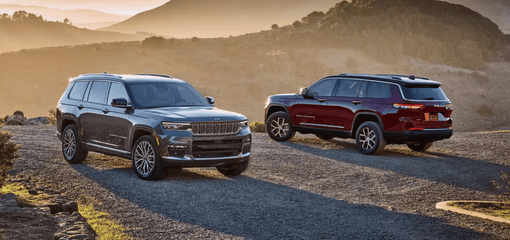A 2025 Jeep Grand Cherokee and Grand Cherokee L parked on a scenic mountain road, surrounded by lush greenery and rocky terrain.