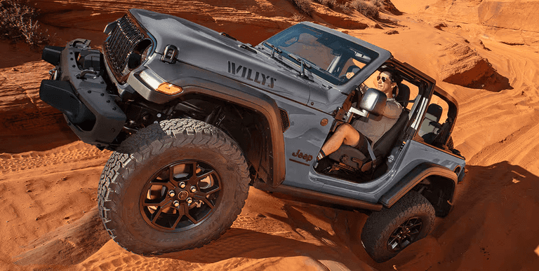 A jeep wrangler traverses a vast desert landscape, kicking up sand as it moves through the arid terrain under a clear sky.