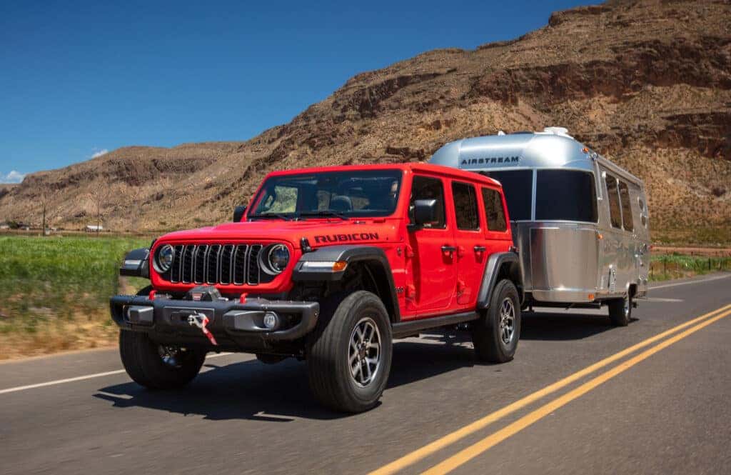 A red 2025 Jeep Rubicon towing a recreational vehicle on a scenic road, showcasing a perfect outdoor adventure setup.