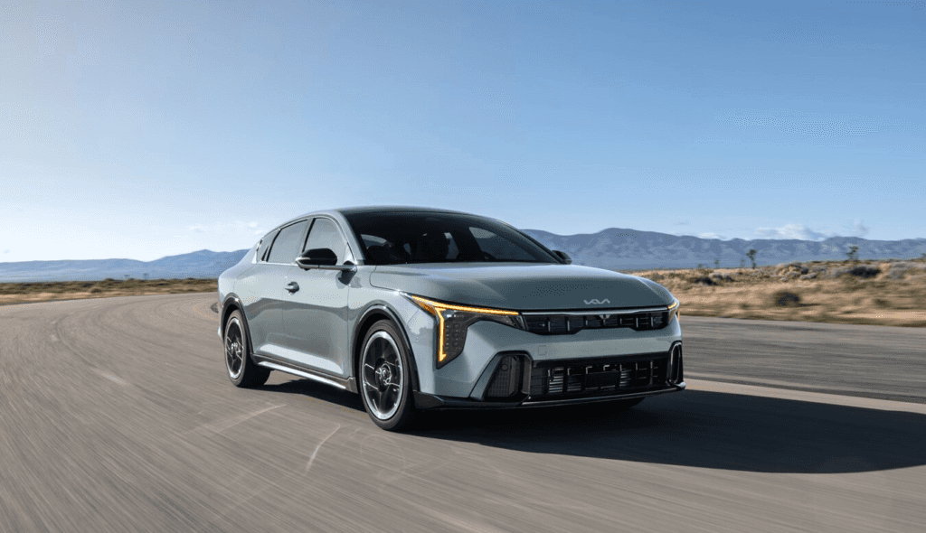 A 2025 KIA k4 compact sedan navigates a sunlit desert road, showcasing its sleek design against a backdrop of sand dunes.