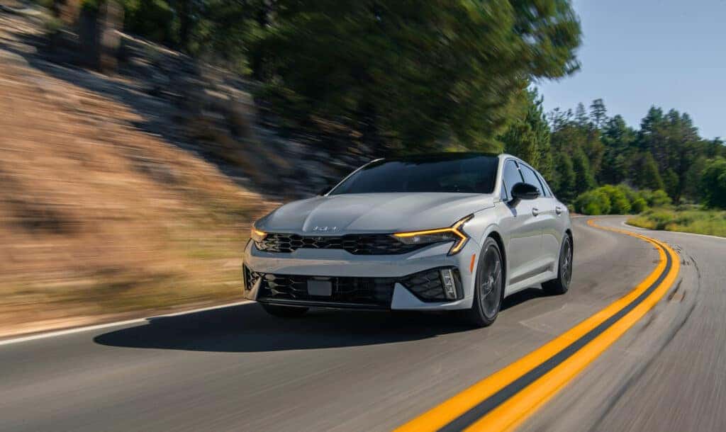 A 2025 Kia K5 navigates a winding mountain road, showcasing its sleek design against a scenic backdrop.